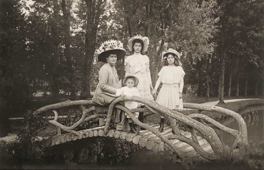 women with victorian flowered hats standing on a faux bois bridge